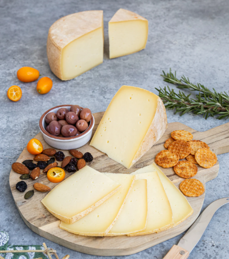 A styled cheese charcuterie setup on a round wooden board, featuring several wedges and slices of pale yellow cheese, almonds, dried fruits, chocolate-covered olives in a small bowl, round crackers, kalamite oranges, and rosemary sprigs on a gray stone surface.